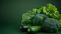 Fresh green vegetables including broccoli, zucchini and lettuce leaves arranged on dark green background with copy space for healthy eating concept.