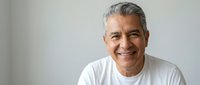 Confident mature Hispanic man with gray hair smiling warmly in white t-shirt against neutral background for lifestyle and wellness content.
