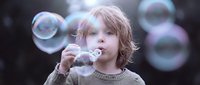 Young blonde child blowing soap bubbles outdoors with colorful iridescent spheres floating around creating magical childhood moment for family content.