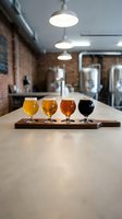 Beer flight tasting sampler with four different craft brews on wooden board in modern brewery taproom with industrial lighting and brewing equipment.