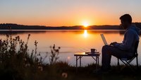 Young professional man working remotely on laptop by serene lake during golden sunset, enjoying work-life balance and digital nomad lifestyle outdoors.