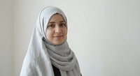 Young Middle Eastern woman wearing gray hijab smiling confidently in modern office setting for professional business and career concepts.