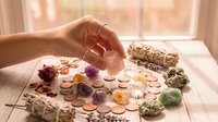 Female hand selecting healing crystals from collection on wooden table with sage bundles and candles for spiritual wellness meditation practice.