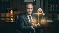 Professional mature Caucasian male lawyer in dark suit holding golden scales of justice in elegant law office library with legal books and warm lighting.