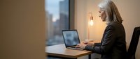 Mature woman with white hair, Caucasian ethnicity, wearing professional black blazer, working on laptop at desk by window in modern office setting during evening hours.