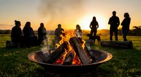 Group of friends gathering around outdoor fire pit during golden hour sunset creating warm social atmosphere for camping and leisure activities.