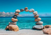 Balanced stones forming arch over turquoise sea water and mountain landscape. Natural zen meditation composition on beach with stacked rocks.