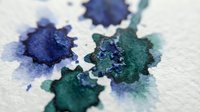 Watercolor paint splatters in deep blue and teal green on white paper, creating organic abstract shapes with soft bleeding edges and textured pigment dispersal.