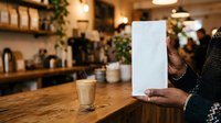 Person holding blank white menu or tablet in modern cafe with wooden counter, warm lighting, and blurred background with plants and shelving for restaurant, hospitality, or business mockup use.
