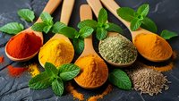 Colorful spices and herbs in wooden spoons on dark slate background. Fresh mint leaves accent turmeric, paprika, cumin and other aromatic seasonings for cooking.