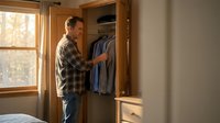 Middle-aged Caucasian man in plaid shirt selecting clothes from wooden wardrobe in cozy bedroom with natural lighting for morning routine concept.