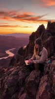 Woman working on laptop at sunset on rocky mountain peak overlooking river valley. Remote work, freelance, digital nomad lifestyle concept for business and travel themes.