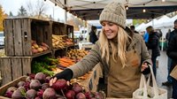 Young Caucasian woman in knit hat shopping at outdoor farmers market, selecting fresh beets among colorful autumn vegetables.