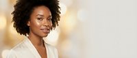 Professional African American businesswoman with curly hair in white blazer smiling confidently against warm bokeh background for corporate presentations.