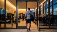 Athletic man entering modern fitness center through glass doors at sunset, ready for evening workout session in well-equipped gym facility.