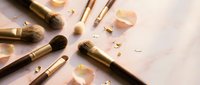 Luxury makeup brushes with gold ferrules scattered on marble surface with rose petals and glitter for beauty and cosmetics branding.