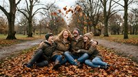 Family of four sitting together on autumn leaves in park, enjoying fall season outdoors with bare trees in background.