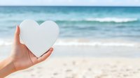 Female hand holding white paper heart shape against blurred ocean beach background with turquoise water and sandy shore, romantic summer concept.