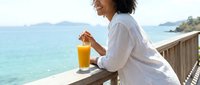 Woman with curly dark hair, mixed ethnicity, relaxing on a coastal deck with orange juice, overlooking turquoise ocean waters and distant islands on a sunny day.