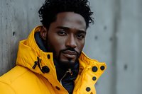 Young African American man wearing bright yellow winter jacket and black turtleneck sweater, looking confidently at camera against concrete wall background.
