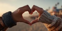 Hands forming heart shape at sunset with warm knitted sweaters creating romantic gesture of love and connection during golden hour outdoors.