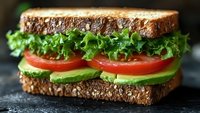 Fresh vegetarian sandwich with lettuce, tomato and avocado on multigrain bread served on dark slate surface for healthy eating and lunch concepts.