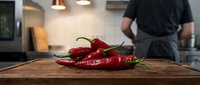 Fresh red chili peppers on wooden cutting board in modern kitchen with chef preparing food in background for culinary cooking and spicy recipe preparation.