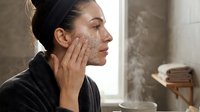 Young woman applying exfoliating face scrub in modern bathroom during morning skincare routine for healthy glowing skin and self-care wellness.