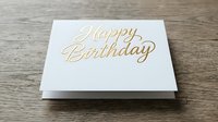 Elegant gold foil Happy Birthday greeting card on white paper placed on rustic wooden surface for celebration and party invitations.