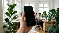 Hand holding smartphone with blank black screen in modern coworking office, green plants and colleagues working in background.