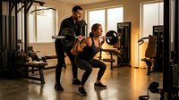 Personal trainer coaching woman during barbell squat exercise in modern gym with natural lighting and professional equipment.