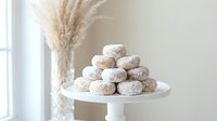 Powdered sugar donuts stacked on white cake stand with pampas grass decoration in bright minimalist kitchen interior for bakery display and food styling.