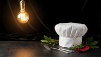 Professional chef hat with fresh herbs and chili pepper on dark wooden surface under warm Edison bulb lighting for culinary restaurant kitchen concepts.