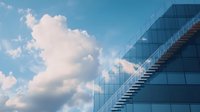 Modern glass building facade with exterior staircase against blue sky with fluffy white clouds, representing corporate architecture and urban development.