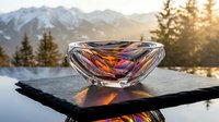 Luxury crystal bowl with colorful light reflections on slate surface, mountain landscape and pine trees in golden sunset background.