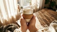 Hands holding ceramic coffee mug in cozy morning sunlight streaming through window. Warm hygge lifestyle moment with natural textures and soft lighting.