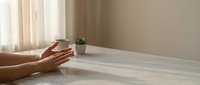 Hands reaching toward small potted plant on wooden floor in minimalist room with natural light streaming through sheer curtains creating peaceful atmosphere.