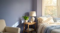 Cozy modern bedroom interior with purple walls, wooden nightstand, table lamp, green plant and comfortable bed with soft lighting from window.