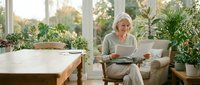 Senior Caucasian woman reading book in bright sunroom surrounded by houseplants, relaxing in comfortable chair by large windows with natural light.