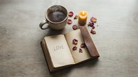 Steaming coffee cup, open vintage book with love inscription, glowing candle and dried rose petals on wooden table, romantic cozy mood.