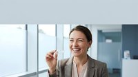 Professional businesswoman in beige suit smiling confidently while holding pen in modern office environment with large windows and natural lighting.