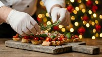 Professional chef in white gloves garnishing festive holiday appetizers with fresh rosemary on wooden board against Christmas tree lights background.