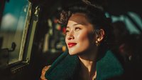 Young Asian woman in vintage 1940s style with victory rolls hairstyle and green coat looking pensively out train window during golden hour journey.