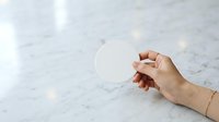 Hand holding blank white circle on marble surface with soft lighting for branding mockup and product presentation purposes.