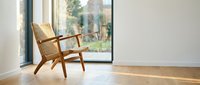 Wooden lounge chair positioned by large window in minimalist interior space with natural light, hardwood flooring, and garden view for modern home design and furniture styling.