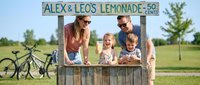 Family running lemonade stand outdoors on sunny day. Woman and man with two young children selling refreshments at wooden booth with sign. Summer activity, entrepreneurship, and childhood memories.