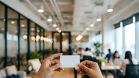 Professional holding blank white business card in modern office environment with colleagues working in background and natural light from windows.