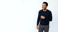Young man with dark hair and beard, South Asian ethnicity, wearing navy blue sweater and gray pants, smiling and gesturing while speaking against white background for advertising and promotional use.