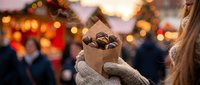 Woman holding paper bag of roasted chestnuts at festive Christmas market with warm bokeh lights creating cozy winter atmosphere.