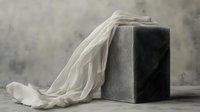Minimalist still life with flowing white silk fabric draped over gray concrete cube against moody atmospheric background. Fine art composition for interior decor.